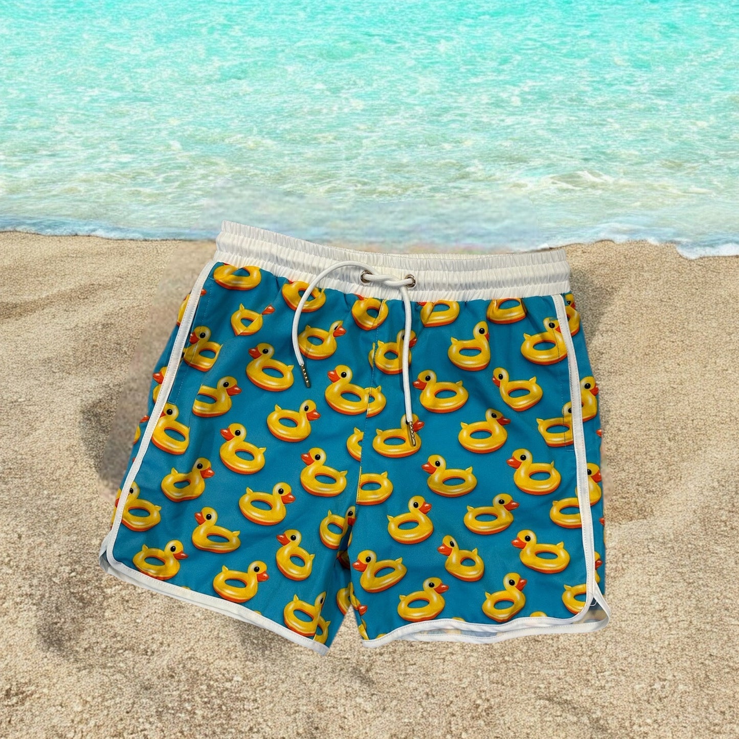 Boys swim shorts with rubber ducky fun print, displayed on a beach background.