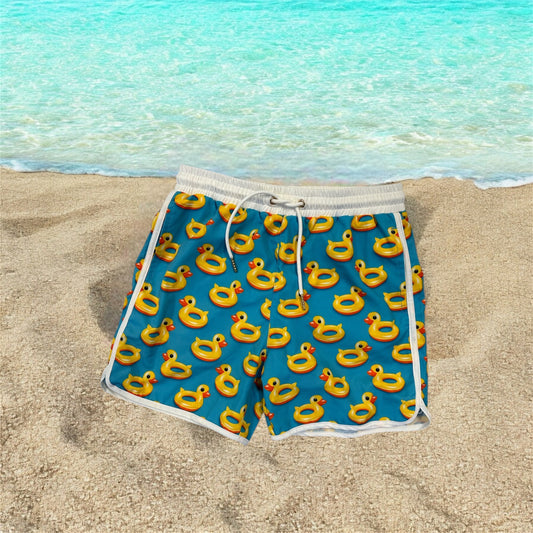 Boys swim shorts with rubber ducky fun print, displayed on a beach background.